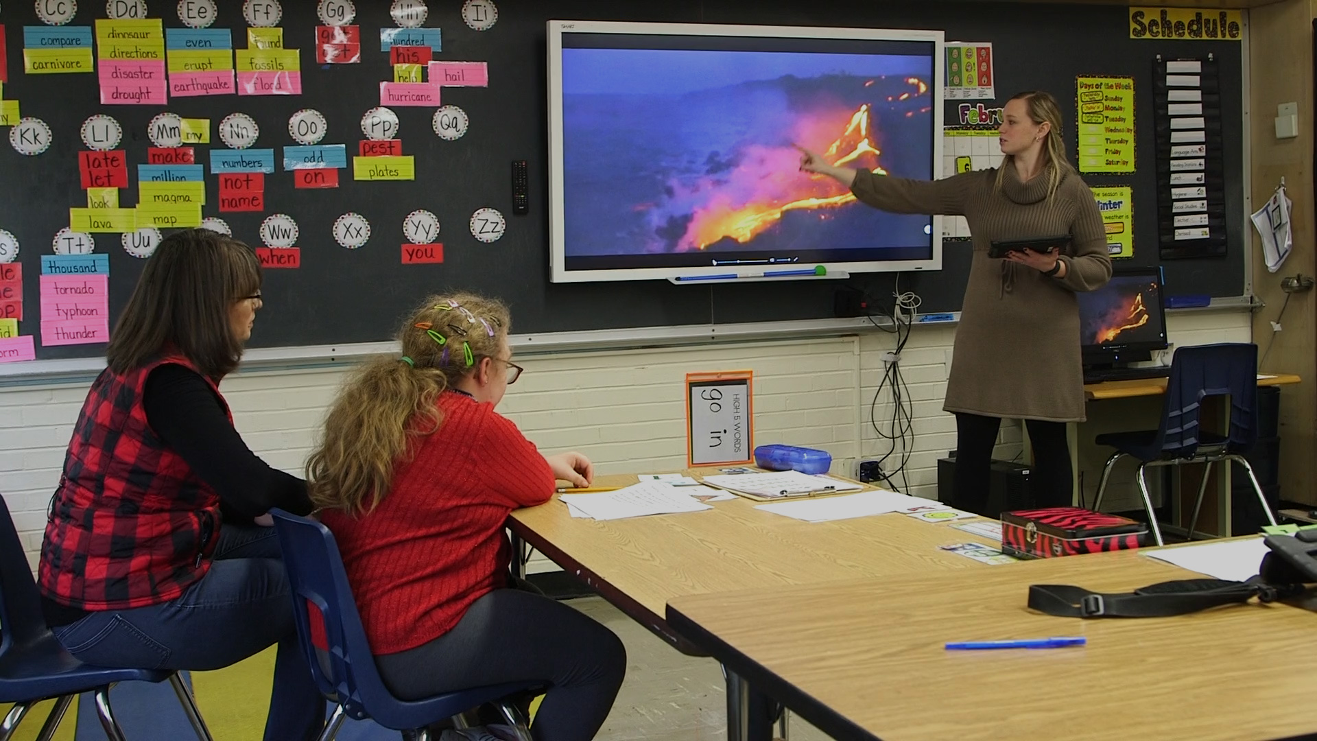 Teacher addresses pupils in front of classroom with TV screen showing volcano erupting