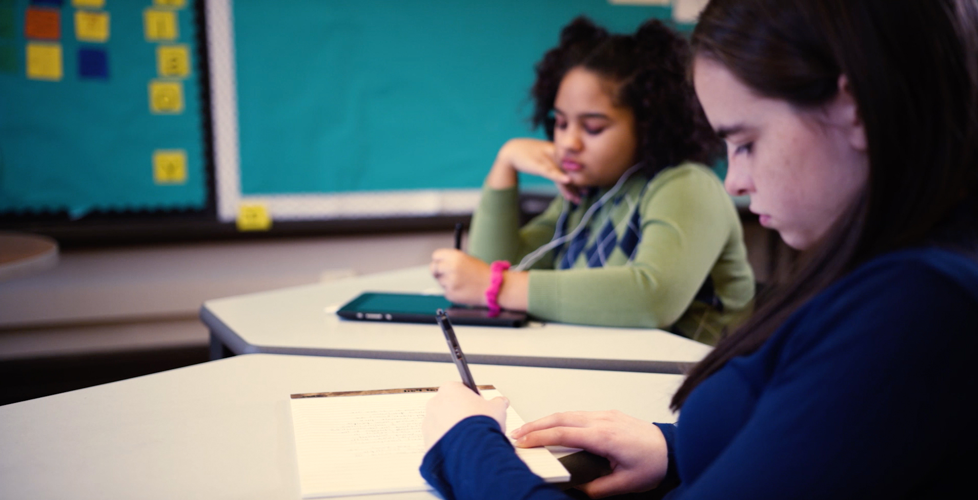 Two students write at desks, one on a tablet, the other in a notebook